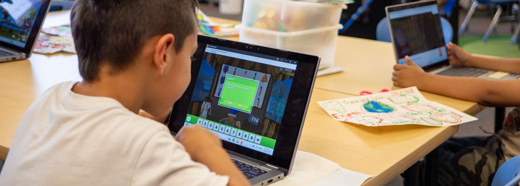 A young student typing on a laptop in a colorful classroom, surrounded by books and learning materials.