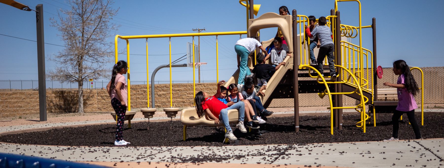 Students happily play on a colorful playground structure, climbing and sliding down the slide.