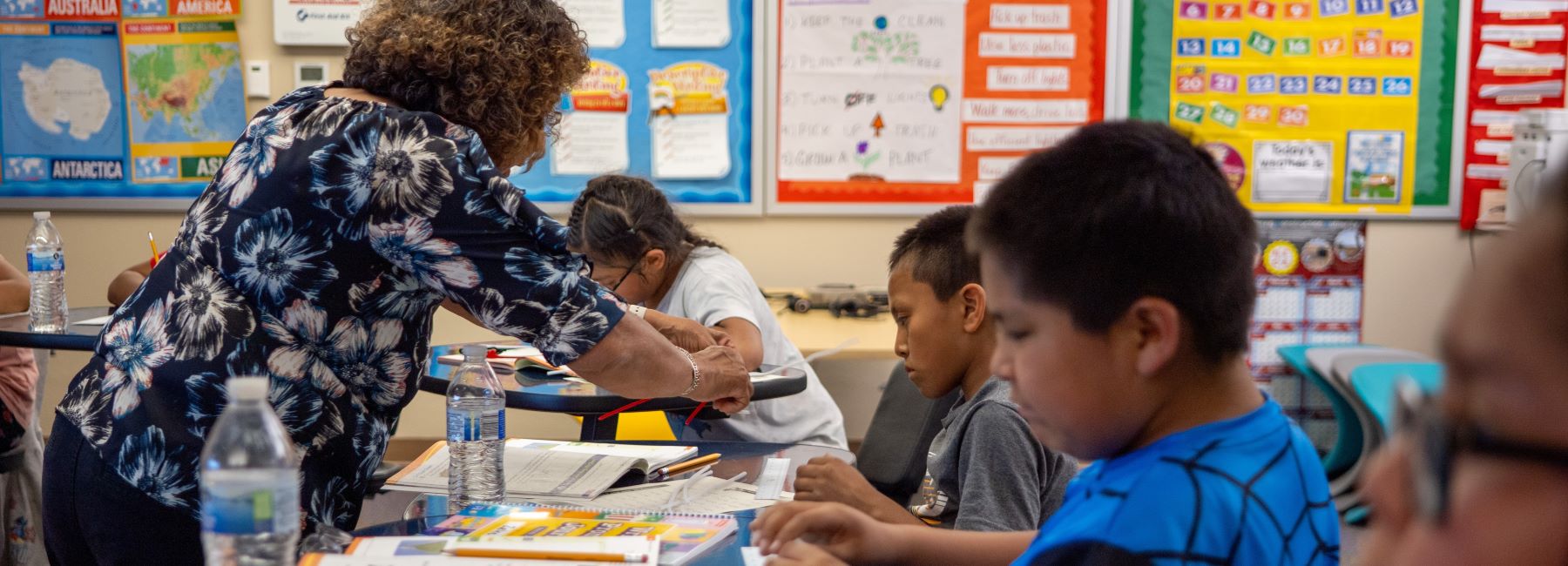 A teacher helps a student in class, leaning over a desk while other student work on their assignments..